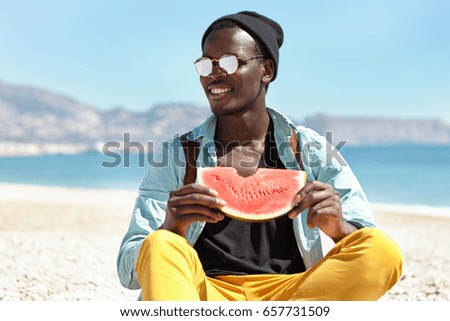 Stylish African treveler in blue shirt, yellow trousers and sunglasses sitting at beach eatting watermelon having wonderful day near sea. Tourist male admiring sea landscape while having rest