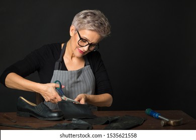 Stylish Aged Woman In Glasses On A Black Background Cuts Leather Material For Shoes