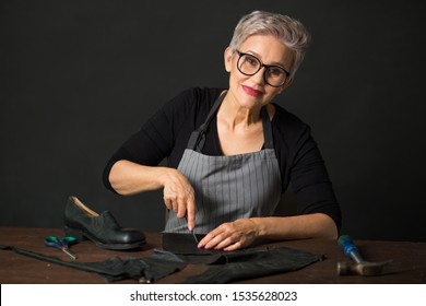 Stylish Aged Woman In Glasses On A Black Background Cuts Leather Material For Shoes