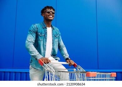 stylish American man wearing sunglasses, running with shopping cart and smiling at camera. A handsome man is shopping in the city and smiling while walking near the mall. Shopping, consumerism concept