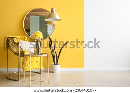 Stylish armchair, vase with blooming narcissus flowers on coffee table and mirror near yellow wall