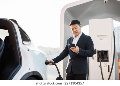 Stylish asian employer with smartphone in hand standing near charging electric car outdoors. Confident man in formal attire using modern app for auto at city power station. - Powered by Shutterstock