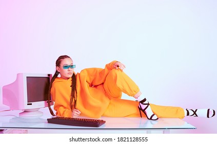 Stylish Asian Female Office Worker In Sunglasses Laying On Table With Obsolete Computer. Colored Neon Light