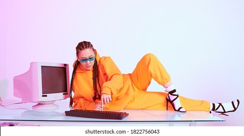 Stylish Asian Female Office Worker In Sunglasses Laying On Table With Obsolete Computer Typing In Neon Light