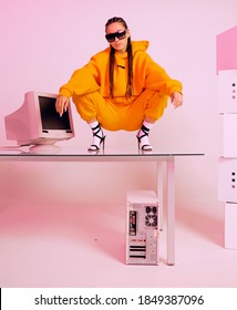 Stylish Asian Female Office Worker With Obsolete Computer Squats On Desk. Colored Neon Light