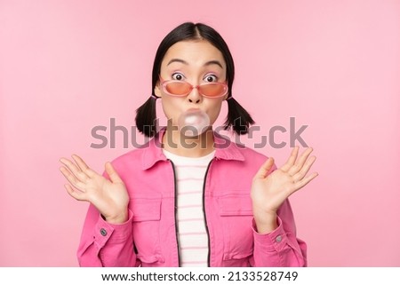 Stylish asian girl blowing bubblegum bubble, chewing gum, wearing sunglasses, posing against pink background