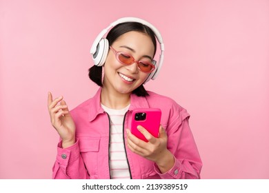 Stylish asian girl dancing with smartphone, listening music in headphones on mobile phone app, smiling and laughing, posing against pink background