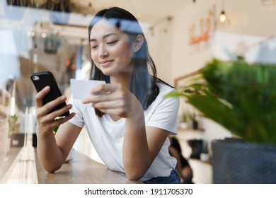 Stylish Asian Girl Sitting In Cafe Near Window, Paying For Online Shopping With Credit Card, Transfer Money For Coffee With Smartphone, Casually Sitting In Restaurant On Summer Day.