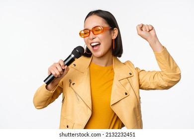 Stylish asian girl in sunglasses, singing songs with microphone, holding mic and dancing at karaoke, posing against white background