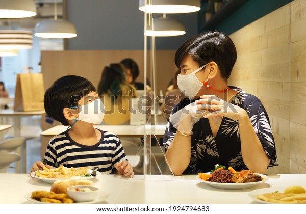 Stylish Asian mother and her child wear face\
mask, smile and look at each other through clear acrylic table\
barrier that separate them apart in beautiful food court. New\
normal and social\
distancing.
