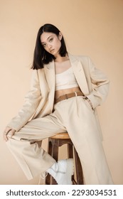 stylish asian woman in pantsuit holding hand in pocket while posing on stool isolated on beige