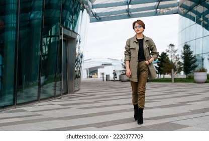 Stylish Asian Woman Wearing Green Suit And Fashion Glasses Walking In Business District While Looking At Camera. Young Woman With Short Haircut Walking Down Street Outdoors.