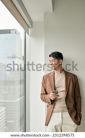 Stylish Asian young business man leader holding phone looking at window standing in modern office. Japanese male professional company executive using smartphone thinking of business vision. Vertical