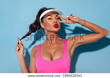 Stylish attractive girl with black hair, bright lipstick, white manicure and earrings in pink bathing suit showing peace sign..