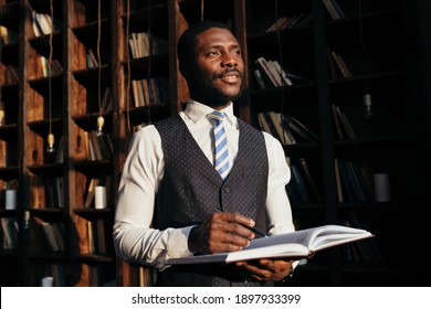Stylish attractive male African American teacher at the university stands with a magazine on the background of shelves with books