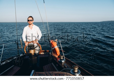A stylish attractive man stands at the helm of the yacht on which he floats on the sea at sunset
