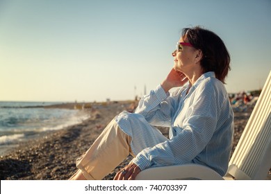 Stylish Attractive Mature Woman 50-60 Years Old Sitting In A Deckchair On The Seashore, Traveling And Retirement, Fashion And Beauty