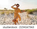 stylish attractive slim smiling woman on beach in summer style fashion trend outfit carefree and happy, feeling freedom, wearing yellow printed dress boho style chic and sunglasses