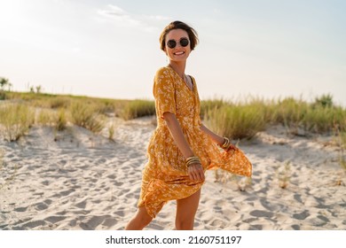 stylish attractive slim smiling woman on beach in summer style fashion trend outfit carefree and happy, feeling freedom, wearing yellow printed dress boho style chic and sunglasses