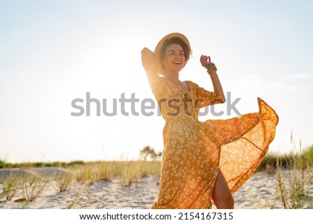 stylish attractive slim smiling woman on beach in summer style fashion trend outfit carefree and happy, feeling freedom, wearing yellow printed dress boho style chic and straw hat