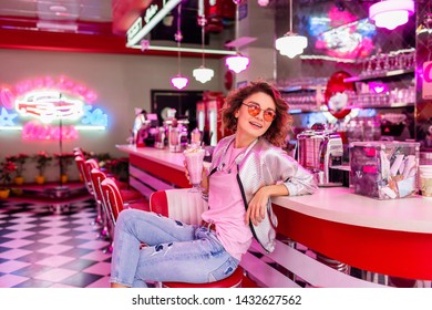 Stylish Attractive Smiling Woman In Retro Vintage Cafe Sitting At Bar Drinking Milk Shake Cocktail In Hipster Outfit Having Fun Laughing In Cheerful Mood, Wearing Pink Sunglasses, Silver Bomber Jacket