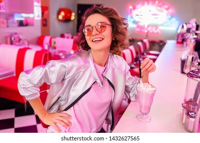 Stylish Attractive Smiling Woman In Retro Vintage Cafe Sitting At Bar Drinking Milk Shake Cocktail In Hipster Outfit Having Fun Laughing In Cheerful Mood, Wearing Pink Sunglasses, Silver Bomber Jacket