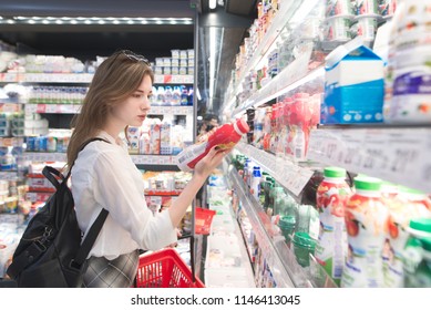 Stylish Attractive Woman Is Standing By The Refrigerator With Dairy Products, Holds Yogurt In His Hands And Reads The Label. An Attractive Girl Buys A Yogurt In A Supermarket.