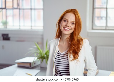 Stylish attractive young redhead businesswoman with a lovely smile standing in front of a table in the office grinning at the camera