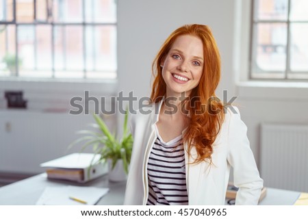 Stylish attractive young redhead businesswoman with a lovely smile standing in front of a table in the office grinning at the camera