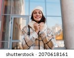 stylish attractive young smiling woman walking in street in winter outfit with coffee wearing checkered coat, white knitted hat and scarf, happy mood, fashion style trend