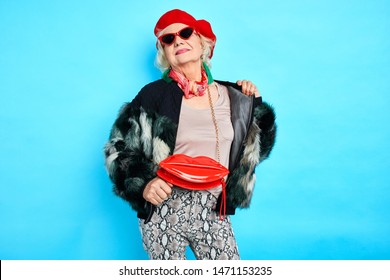 Stylish Awesome Cool Old Woman In Red Hat And Trendy Coat Posing To The Camera. Isolated Blue Background. Studio Shot.autumn, Fall, Spring Clothes . Stylish Clothes For Any Age