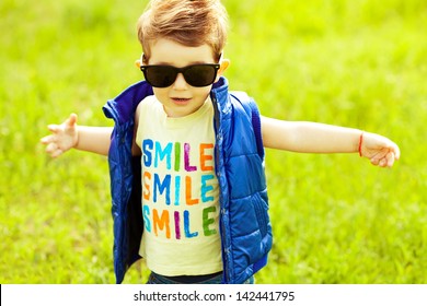 Stylish baby boy with ginger (red) hair in trendy sunglasses and blue jacket standing in the park & trying to hug somebody. Hipster style. Sunny weather. SMILE word printed on t-shirt. Outdoor shot