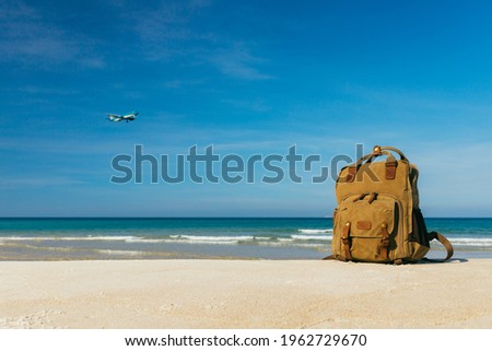 Stylish backpack on a snow-white empty beach by the sea, with an airplane in the background. Travel concept on the lung. Calm waves on the sea, blue clear sky