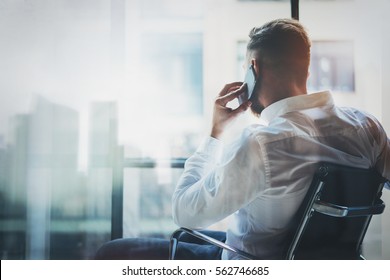 Stylish Banker Making Mobile Call At Modern Loft After Work Day.Man Sitting In Chair And Looking Out The Panoramic Window.Skyscraper Office Building Blurred On Background.Horizontal