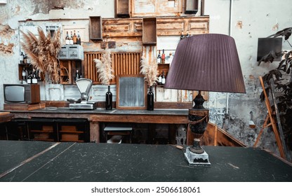 A stylish bar counter featuring a vintage table lamp with a pleated shade in the foreground. The background showcases rustic decor with wooden shelves, a wine rack, and pampas grass in vases - Powered by Shutterstock