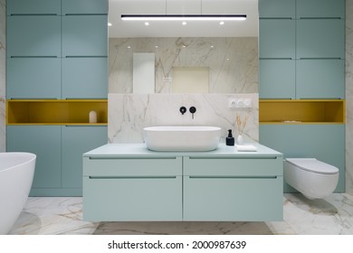 Stylish bathroom with white washbasin with black tap in modern blue chest of drawers and blue cabinets with golden shelves