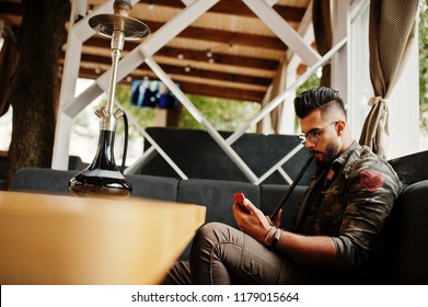 Stylish Beard Arabian Man In Glasses And Military Jacket Smoking Hookah At Street Bar. Arab Model Having Rest And Looking On Phone.