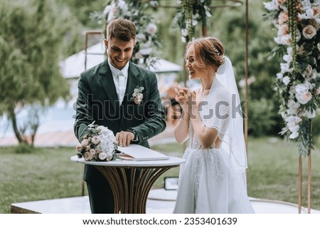 Stylish bearded bride and groom in a white dress are smiling while standing at the ceremony against the backdrop of a decorated arch. Wedding photography, portrait of the newlyweds.