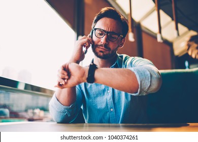 Stylish Bearded Businessman 30 Years Old Looking At Smartwatch While Solving Financial Promlems During Mobile Conversation.Male Entrepreneur Talking On Smartphone About Meeting With Partner In Cafe