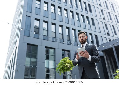 Stylish Bearded Businessman In Formal Business Suit Standing Working With Tablet In Hands On Background Modern Office Building Outside. Man Using Smartphone Or Uses Mobile Phone Outdoors City Street