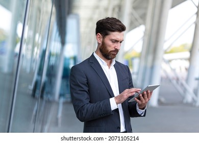 Stylish Bearded Businessman In Formal Business Suit Standing Working With Tablet In Hands On Background Modern Office Building Outside. Man Using Smartphone Or Uses Mobile Phone Outdoors City Street