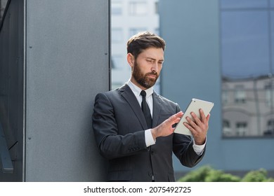 Stylish Bearded Businessman In Formal Business Suit Standing Working With Tablet In Hands On Background Modern Office Building Outside. Man Using Smartphone Or Uses Mobile Phone Outdoors City Street