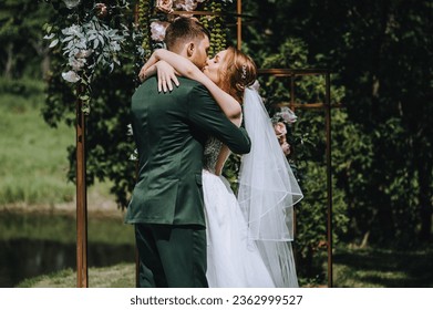 A stylish bearded groom and a beautiful bride in a white dress are hugging, kissing while standing at a ceremony against the backdrop of a decorated arch. Wedding photography, portrait. - Powered by Shutterstock