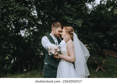 A stylish bearded groom in a suit and a bride in a white dress gently hug while standing in a park in nature with trees. Wedding photography, portrait of the newlyweds. - Powered by Shutterstock