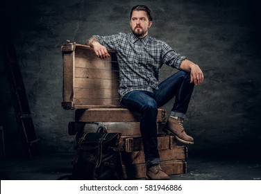 The stylish bearded hipster male sits on a wooden box in a studio over dark grey background.