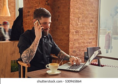 Stylish Bearded Hipster Male Talking By Smartphone And Using A Laptop In A Coffee Shop.