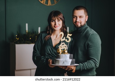 Stylish bearded man and beautiful brunette girl hold a delicious birthday cake with numbers 25 in the interior. Festive photo.