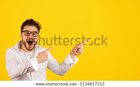Stylish bearded man in shirt and glasses pointing away with amazement while showing new offer on yellow background
