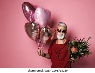 Stylish bearded middle-aged man with a modern hairstyle and fashionably dressed holding bunch of heart shaped balloons and a bouquet of flowers. Concept Valentine's Day, Date - Shutterstock ID 1296735139