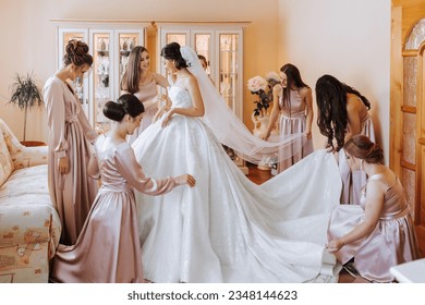 Hermosas damas de honor con vestidos de seda a juego que ayudan a la bella morena novia con vestido blanco a prepararse para la boda, los preparativos de la mañana, la mujer vestida